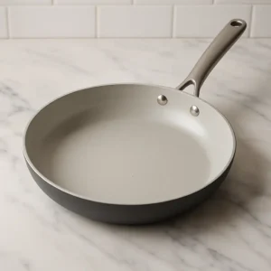 A non-stick ceramic frying pan with a light gray speckled interior and a matte dark gray exterior, resting on a white marble countertop with a stainless steel handle and a white subway tile backsplash.