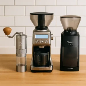 Three coffee grinders displayed side by side on a light wooden countertop in front of a white subway tile wall, including a manual grinder with a wooden handle, a digital Breville grinder with a display screen, and a sleek black Baratza grinder.