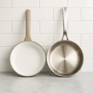 Two frying pans placed side by side on a gray marble countertop with a white subway tile background, one with a white ceramic interior and beige handle, the other made of polished stainless steel.