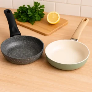 Two frying pans placed side by side on a light wooden countertop — one with a speckled granite surface and black handle, the other with a smooth ceramic coating and beige handle, with a cutting board, parsley, and half a lemon in the background.