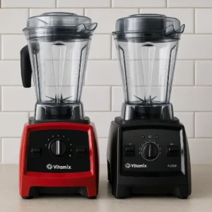 A red Vitamix A2300 and a black Vitamix A2500 blender side by side on a modern kitchen counter with a white tile backsplash.