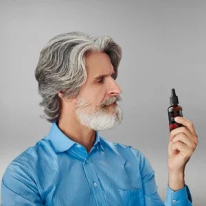 A mature man with gray hair and beard wearing a blue shirt, holding and examining a small dropper bottle, possibly a supplement like Sugar Defender.