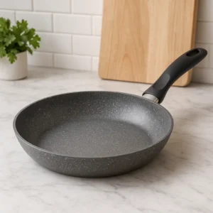 A granite-coated ceramic frying pan on a marble countertop with a cutting board and parsley in the background.